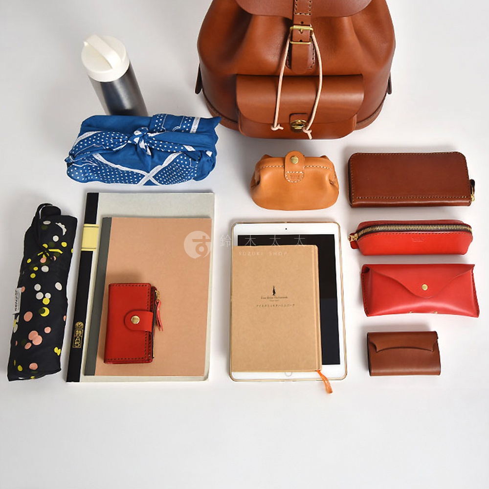 駝色皮革後背包，搭配藍色圓點圖案的折疊傘、白色水壺、藍色印花頭巾、焦糖色小皮夾、棕色長夾、紅色筆袋、紅色信封式皮夾、紅色小卡夾，以及兩本筆記本和一台平板電腦。