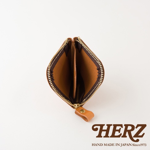 一個呈心形開口的日本HERZ職人手作皮革分隔收納零錢包，選用棕色皮革材質，頂部配有金屬拉鍊與皮革拉環，包內設有分隔層，整體設計簡約精緻並印有HERZ HAND MADE IN JAPAN Since1973字樣。