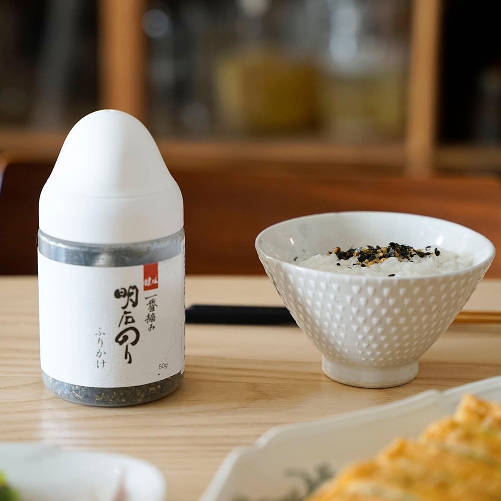 一個白色瓶身、標籤上有日文「一番摘み」及「明太子」字樣的芝麻香鬆，瓶內可見黑白芝麻與碎海苔，旁邊擺著一碗撒有芝麻香鬆的白飯。