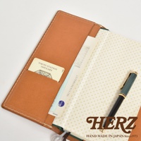 職人手作皮革旅行記事本書套，採用棕色皮革製作，內含卡片夾層，可放置卡片、機票等物品。書套內頁印有規則的圖案，搭配一支綠色筆，展現簡約質感。