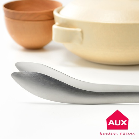 一雙銀色金屬材質、帶有曲型設計的分菜夾，夾頭部分呈現光滑的弧度，背景為淺黃色帶蓋的陶鍋和木色碗。