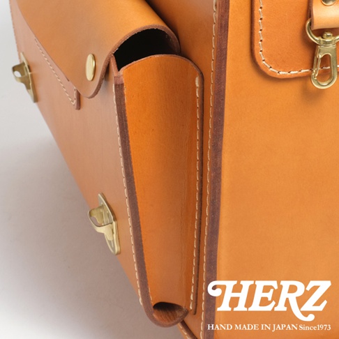 HERZ 品牌標誌，下方為 HAND MADE IN JAPAN Since1973 字樣，搭配一個淺棕色皮革橫式包包的局部特寫，包包上有金屬鎖扣和縫線細節。