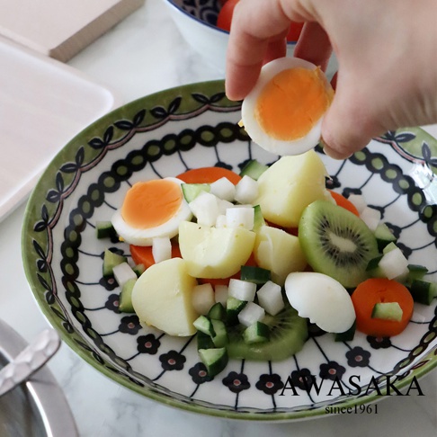 一個裝有水煮蛋、馬鈴薯、小黃瓜、胡蘿蔔和奇異果的陶製料理盤，盤緣飾有綠色葉紋和黑色花朵圖案。