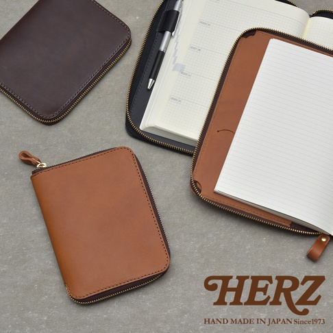 HERZ 品牌標誌，下方為 HAND MADE IN JAPAN Since 1973 字樣。圖片中有三個皮革製品，一個深棕色拉鍊式筆記本收納包，一個淺棕色拉鍊式筆記本收納包，以及一個打開的黑色拉鍊式筆記本收納包，內含一本筆記本和一支筆。