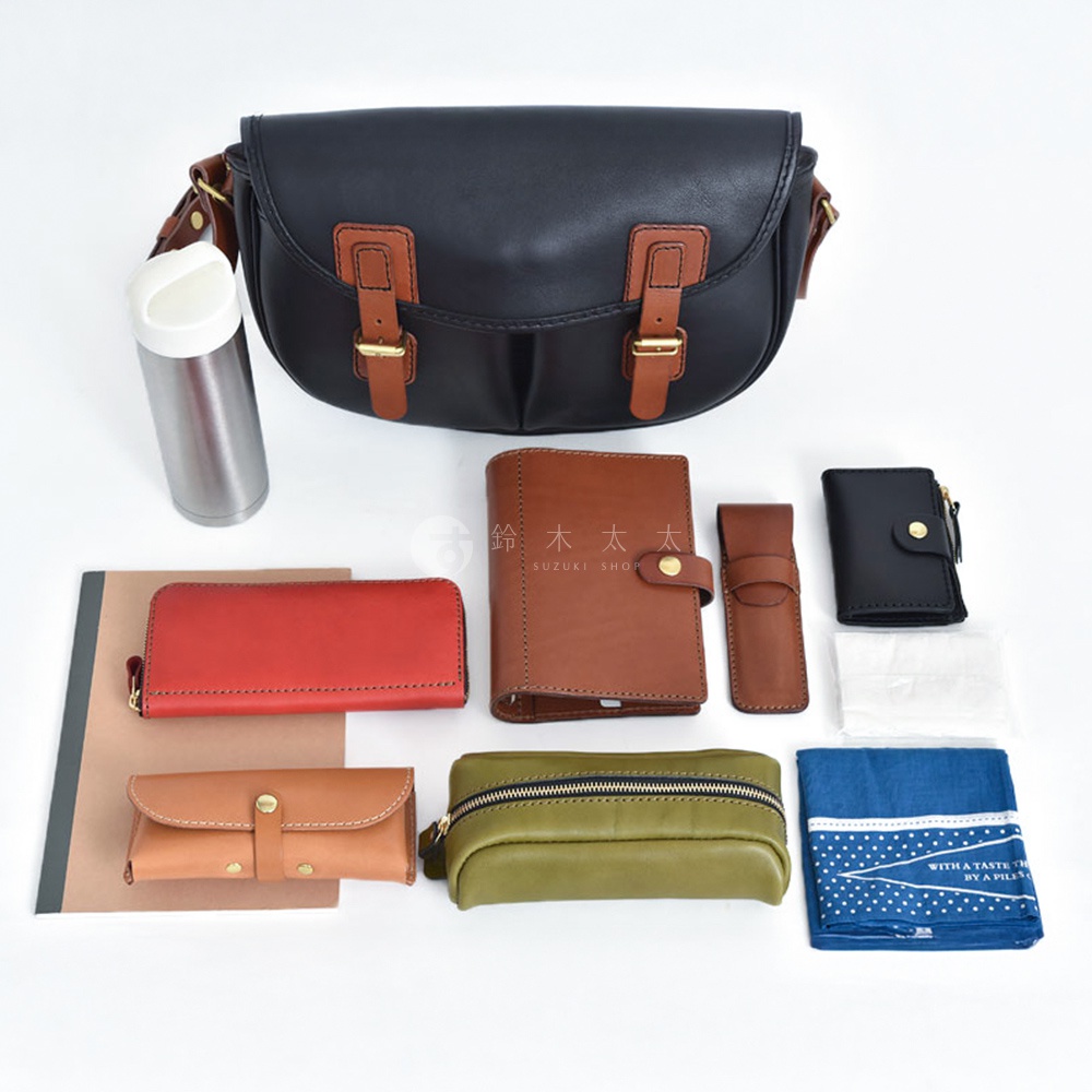 黑色皮革水桶包，搭配棕色皮革飾邊和搭扣。旁邊展示了紅色長夾、棕色筆記本、棕色筆套、黑色零錢包、橄欖綠化妝包、一個銀色保溫瓶和藍色印花手帕。