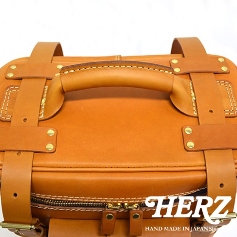 職人手作皮革大型三用旅行後背包，展示其圓潤的頂部提把、兩側的皮革固定帶，以及底部雙拉鍊設計。整體為溫暖的棕褐色，材質為皮革，細節處可見精緻的縫線與金屬卯釘。