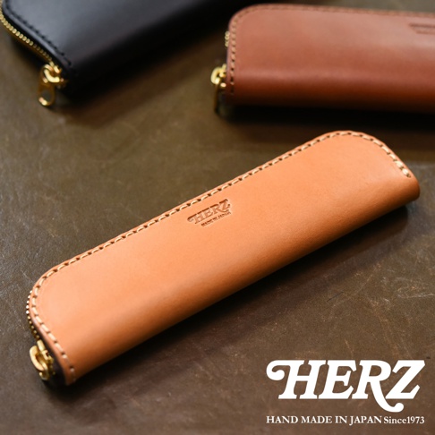 HERZ 品牌壓印的皮革筆袋，淺棕色皮革搭配金色拉鍊，縫線精緻，背景為深棕色皮革桌面。
