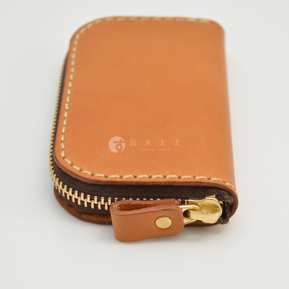 職人手作皮革鑰匙包，橢圓形設計，附有金色拉鍊及皮革拉環，縫線精緻，顏色為淺棕色。