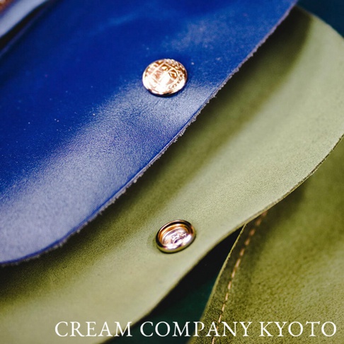 藍色皮革半月形迷你手提包，配有金色按扣，下方為綠色麂皮材質，可見縫線細節。
