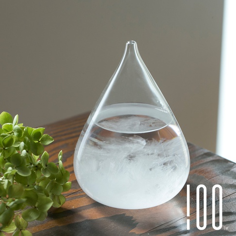 透明玻璃製成的小型水滴狀天氣瓶，內部裝有液體和細小的白色結晶，瓶口細長，瓶身置於木質桌面，旁有一枝綠色植物。