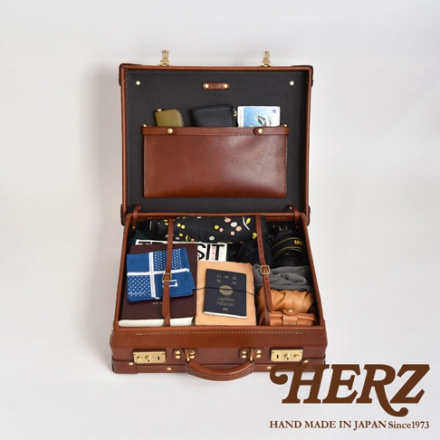 職人手作皮革英倫風便攜手提行李箱，開啟狀態，內部裝有護照、錢包、摺疊傘、相機及衣物等。行李箱為棕色皮革材質，搭配金色鎖扣與提把，內襯為黑色。