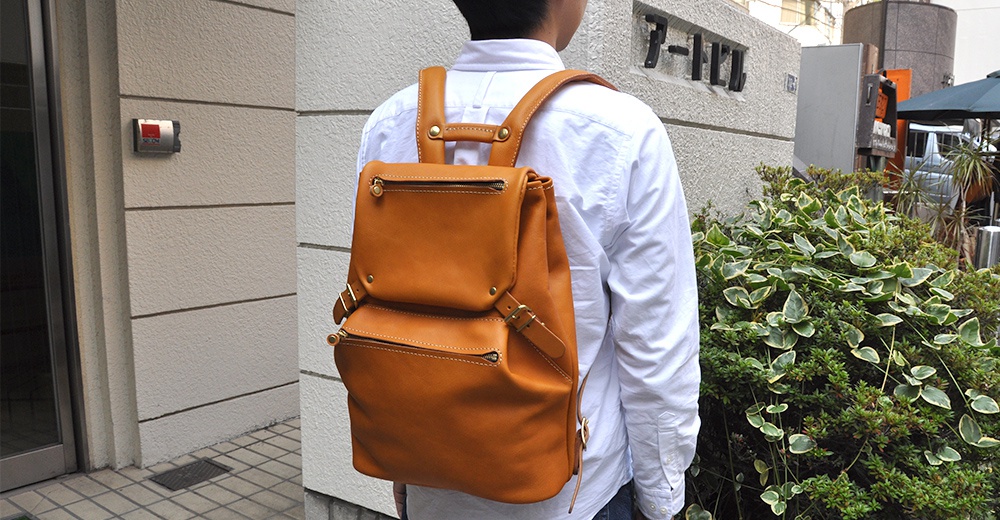 棕色皮革多口袋後背包，具有兩個前置拉鍊口袋和一個翻蓋主袋，配有金屬扣環和可調式肩帶。