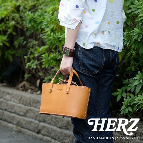 職人手作皮革束口手提包，採用 tan 色皮革製成，簡約的方形設計，附有雙提把，包身印有「HERZ」品牌標誌。