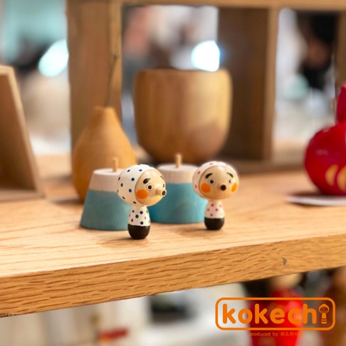 兩尊木芥子開運擺飾，皆為木質，身穿白色頭巾，頭巾上有黑色或紅色圓點圖案，臉部有圓形紅臉頰和黑色眼睛、鼻子。其中一尊穿著黑色圓點上衣，另一尊穿著紅色圓點上衣。背景為淺色木質層架。