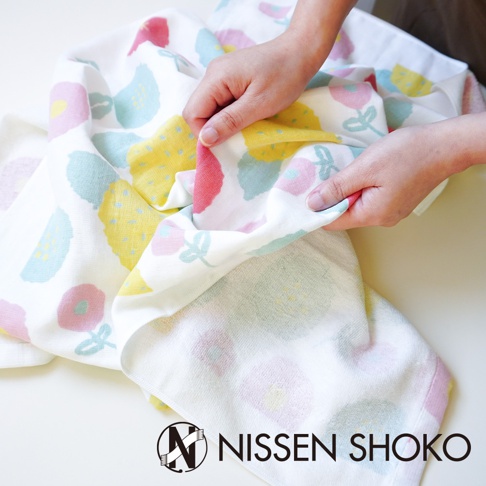 雙手正在展示一條印有粉色、藍色與黃色花草圖案的柔軟棉質浴巾，織紋細緻且觸感舒適，下方印有日纖商工 NISSEN SHOKO 的品牌標誌與名稱。