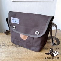 犬印純綿帆布雙扣郵差包，深棕色，配有灰色滾邊和皮革裝飾。包身有兩個銀色旋轉鎖扣，左側有一個印有「犬印製作所」字樣的白色標籤。