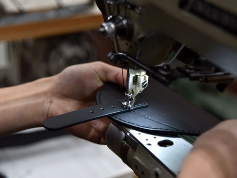 職人以工業縫紉機縫製黑色皮革翻蓋迷你單肩包的皮革部分，展示手作細節。