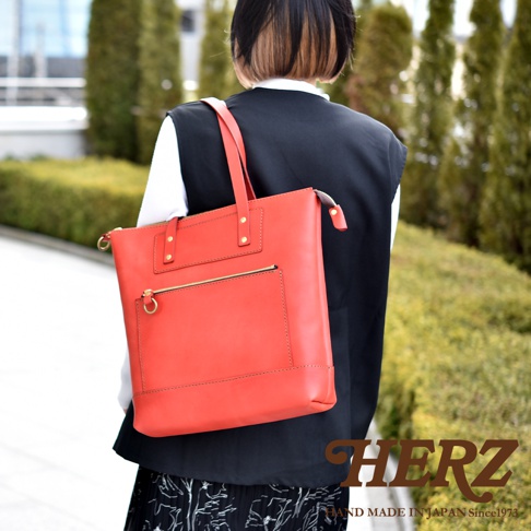 一位短髮女性背著日本HERZ品牌的亮橘色職人手作皮革威靈頓直式拉鍊托特包，包款設計簡約且具有精緻的車縫細節，右下角印有HERZ品牌標誌。