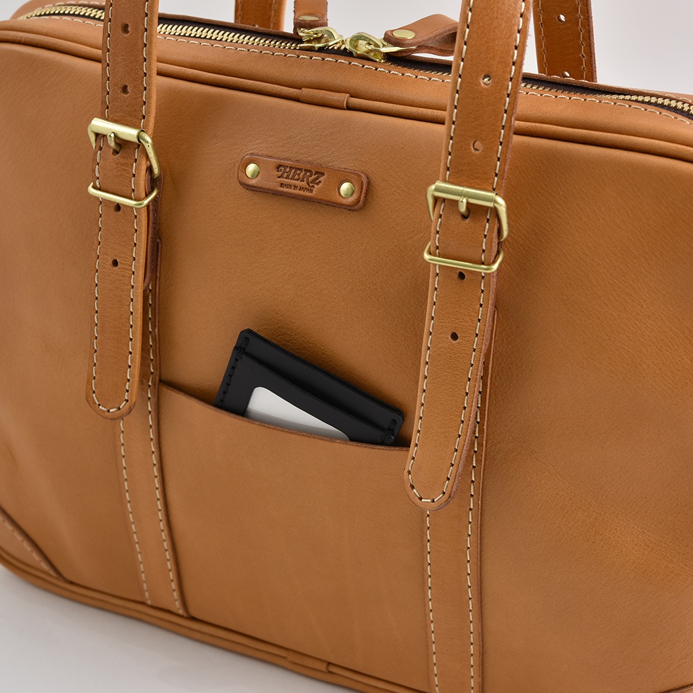 棕色皮革公事包，有兩個可調節的肩帶，配有黃銅扣環。包包正面有一個外袋，內含一個黑色卡套。包包頂部有拉鍊，並設有品牌標誌。