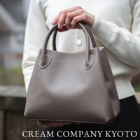 一位女性手持一個灰褐色荔枝紋牛皮托特包，包包有兩個提把，上方有白色縫線裝飾。