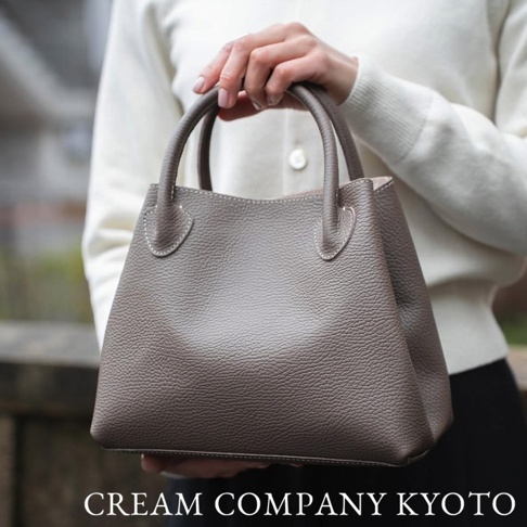 一位女性手持一個灰褐色荔枝紋牛皮托特包，包包有兩個提把，上方有白色縫線裝飾。