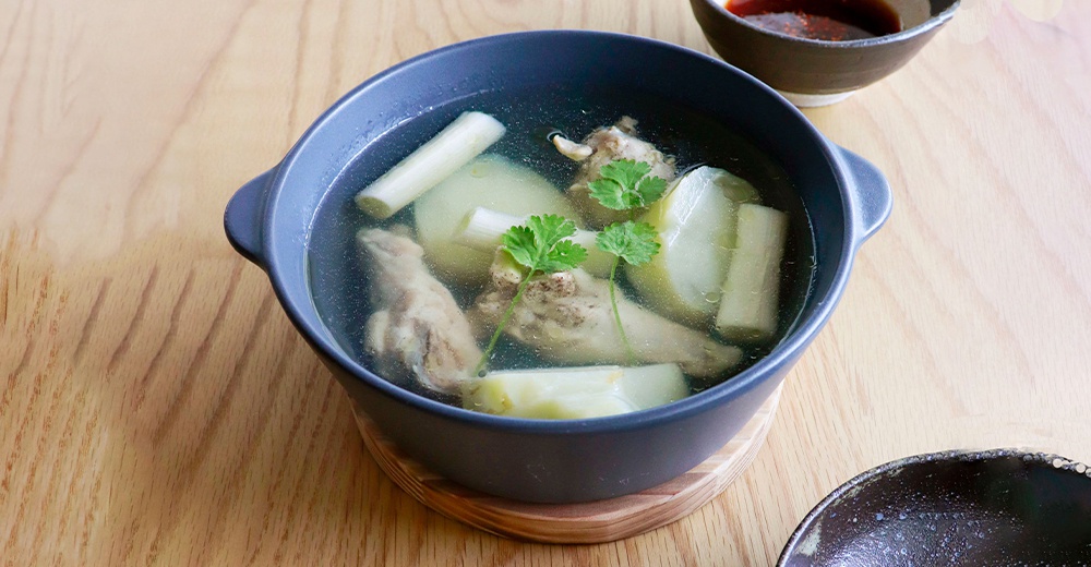 深灰色陶製圓鍋中盛有雞湯，湯內有雞肉塊、冬瓜塊及香菜點綴，鍋具置於木質桌面。