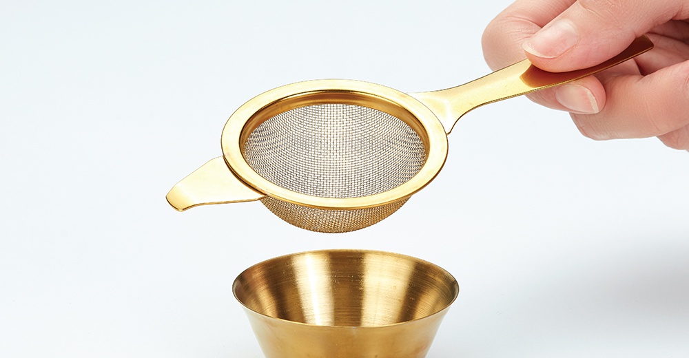 金色金屬製的茶篩，網孔細密，帶有長柄，正懸置於一個同樣是金色金屬製、呈杯狀的容器上方。