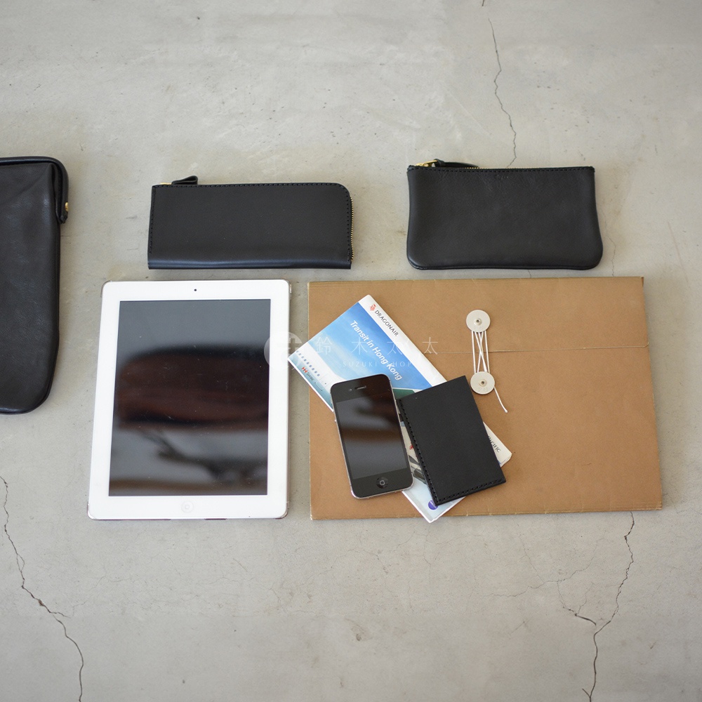 職人手作皮革Organ手拿包 G-38，黑色皮革材質，包含一個長方形拉鍊零錢包，一個較小方形零錢包，以及一個長條形零錢包，旁邊擺放一台白色平板電腦、一本旅遊指南、一支手機及一個黑色卡夾。
