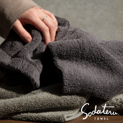 兩條深灰色與淺灰色質地蓬鬆柔軟的浴巾，疊放於桌面，一隻手正觸碰其中一條浴巾。