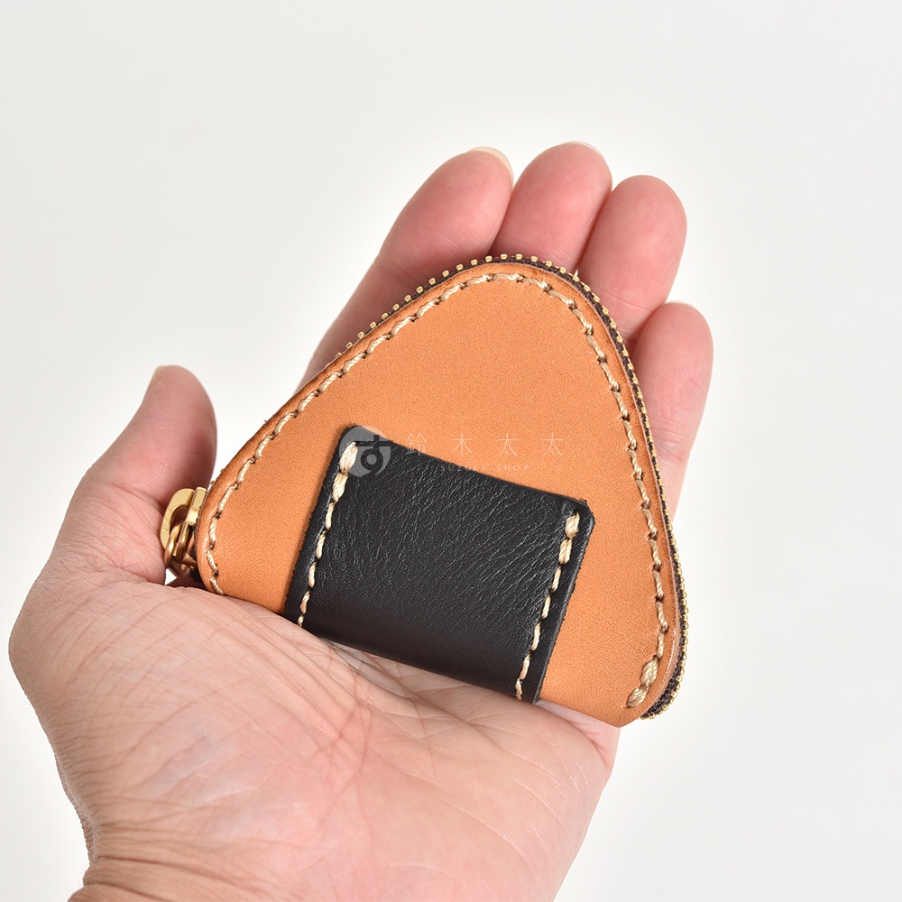 一隻手掌托著一個日本HERZ職人手作皮革三角飯糰造型零錢包，零錢包由淺棕色皮革為主體，搭配黑色皮革細節，邊緣飾有粗線縫製工藝，整體造型精緻小巧。