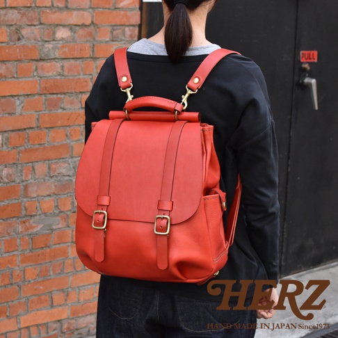 一位女性背著日本HERZ品牌的紅色皮革雙扣後背包，背包上方有堅固的皮革提把，採用經典的屋瓦式頂部結構設計，表面為質感細緻的牛皮材質，畫面右下角呈現HERZ的品牌標誌與手工製造資訊。