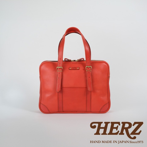 鮮紅色皮革手提肩背兩用公事包，設有雙提把、拉鍊開合，前方有兩個皮革束帶及一個平口袋，搭配金色金屬釦件，下方有皮革加固。