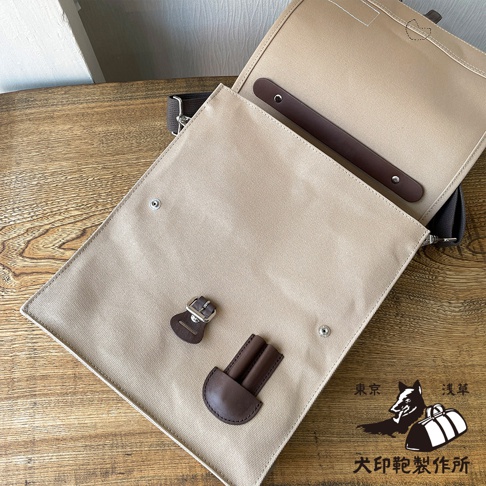 米色純綿帆布製成，直式三用側背包，帶有深棕色皮革飾邊、皮帶扣和皮革插槽。包包開啟，露出內部細節，底部有「犬印鞄製作所」的黑色標誌。