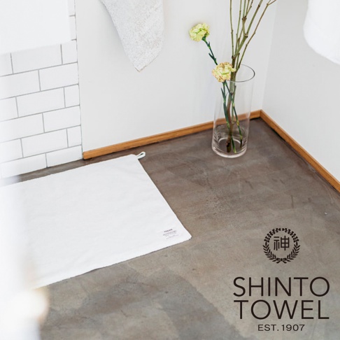 一條鋪在地板上的白色毛巾，旁邊有一盆花和一個寫有 SHINTO TOWEL EST. 1907 的標誌。