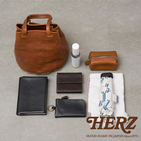 這張由日本HERZ品牌呈現的照片，展示了職人手作皮革Organ抽繩肩背手提托特包及其收納內容物。畫面中央由上方依序為棕色皮革包袋、一瓶噴霧、小型皮革收納袋，下方則整齊擺放著黑色筆記本、深棕色短夾、黑色皮革鑰匙零錢包，以及包裹在淺色條紋摺疊傘旁的白色方巾。整體色調溫潤雅緻，充滿日系職人風格。
