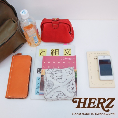 職人手作皮革休閒風後背包 R-93 的配件組，包含一個紅色的皮革零錢包、一個橘色的皮革長夾、一瓶飲料、一本雜誌、一條印有鳥紋的布巾，以及一部白色手機。