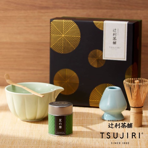 辻利茶舖星之翠茶道具禮盒，包含一個淡綠色瓷製片口碗、一個竹製茶杓、一個淺藍色竹刷架與一個竹製茶筅，背景為帶有金色圓形幾何圖案的黑色包裝禮盒，整組道具陳列於竹編底座上，營造出寧靜雅緻的抹茶品茗氛圍。