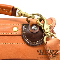 淺棕色皮革郵差包特寫，展示了黃銅色釦環、鹿角扣和細緻的縫線細節。