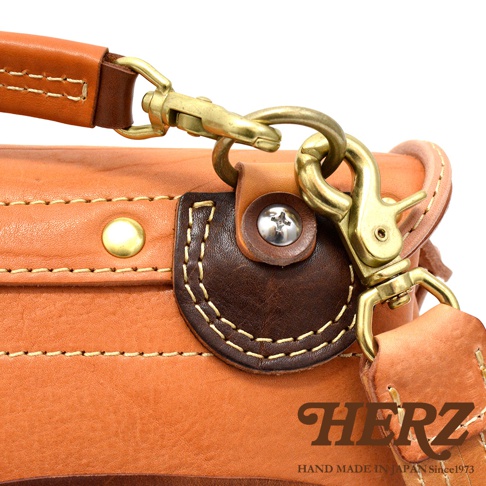 淺棕色皮革郵差包特寫，展示了黃銅色釦環、鹿角扣和細緻的縫線細節。