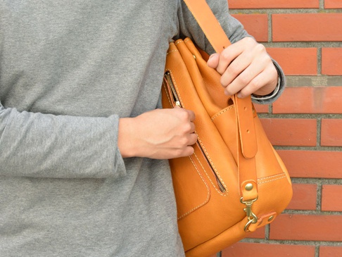 焦糖色皮革雙扣後背包，配有拉鍊口袋及金屬掛鉤，由手部握持，背景為磚牆。
