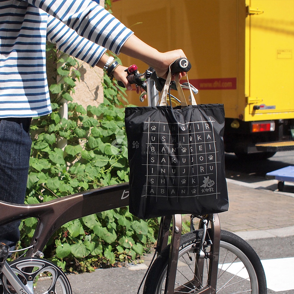一個黑色的環保折疊收納購物袋，正面印有白色網格圖案，其中包含日文文字和狗爪圖案，上方有兩個米白色提把。