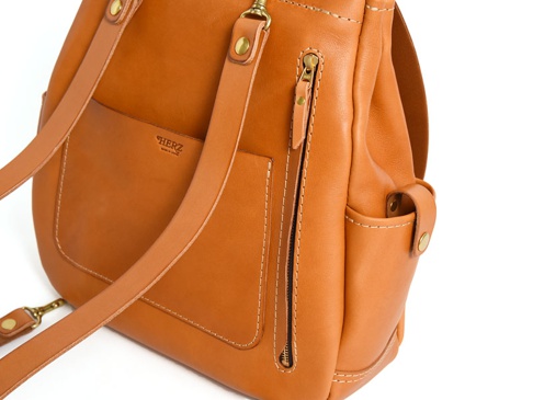 棕色皮革後背包，具有雙扣設計，側面有拉鍊口袋和翻蓋口袋，肩帶採用皮革材質，車縫線明顯。