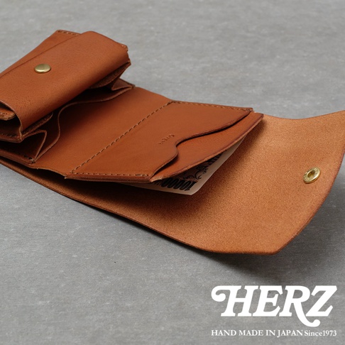 一張展現日本HERZ手工製作的棕色皮革迷你短夾照片，錢包呈現攤開狀態，內側設有卡片收納槽與零錢袋，邊緣可見日本萬元鈔票。整體材質為溫潤的Minerva Box皮革，右下角印有HERZ品牌標誌，下方標註為日本手作，展現精緻細膩的工藝質感。
