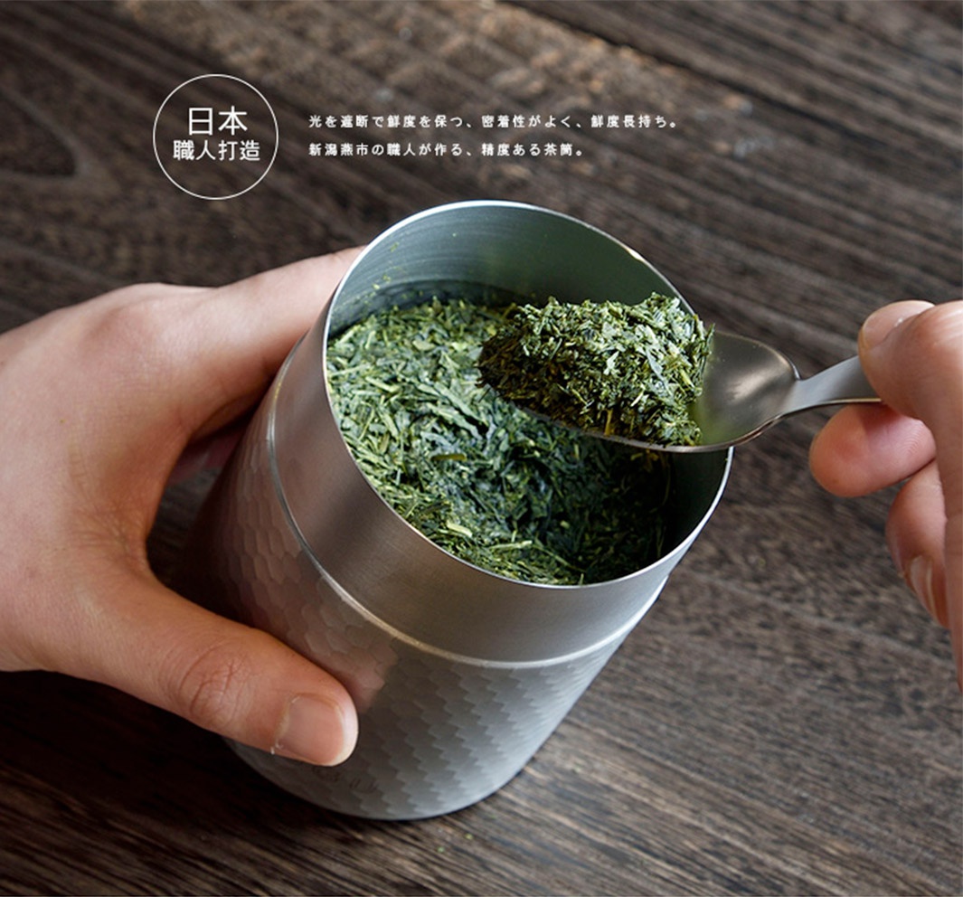 日製18-8不鏽鋼錘目紋茶葉罐，銀色金屬材質，罐身有細緻錘打紋路，罐內裝滿綠色茶葉，附一支金屬茶匙，正在舀取茶葉。