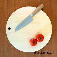 淺色木質圓形砧板，直徑約30公分，側邊有掛孔。上方擺放一把不鏽鋼刀刃、木柄的菜刀，以及兩個切開的紅色番茄。