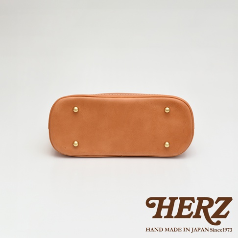 淺棕色皮革貝殼手提包底部，設有四顆金色金屬腳釘，包身以白色縫線裝飾。