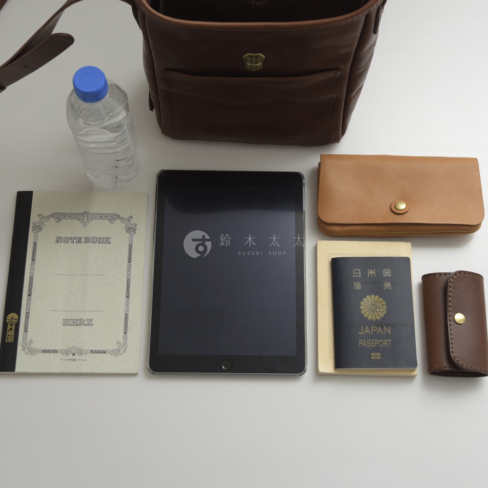 棕色皮革輕便單肩側背包，展示內部空間，擺放著筆記本、平板電腦、皮夾、護照和鑰匙包。