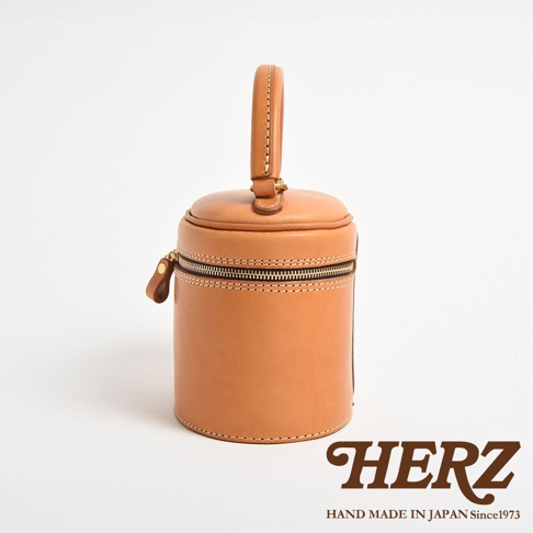 職人手作皮革圓筒型化妝手提包，呈現天然皮革的淺棕色，圓柱形設計，頂部有提把，側邊有拉鍊開合。