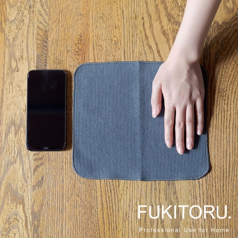 一張深藍色超細纖維清潔擦拭布，置於木紋桌面，旁邊放著一支黑色手機，一隻手放在擦拭布上。