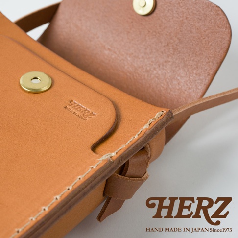 職人手作皮革手機包，材質為棕色皮革，帶有金屬按扣和車縫細節。包身上壓印有「HERZ MADE IN JAPAN」字樣。
